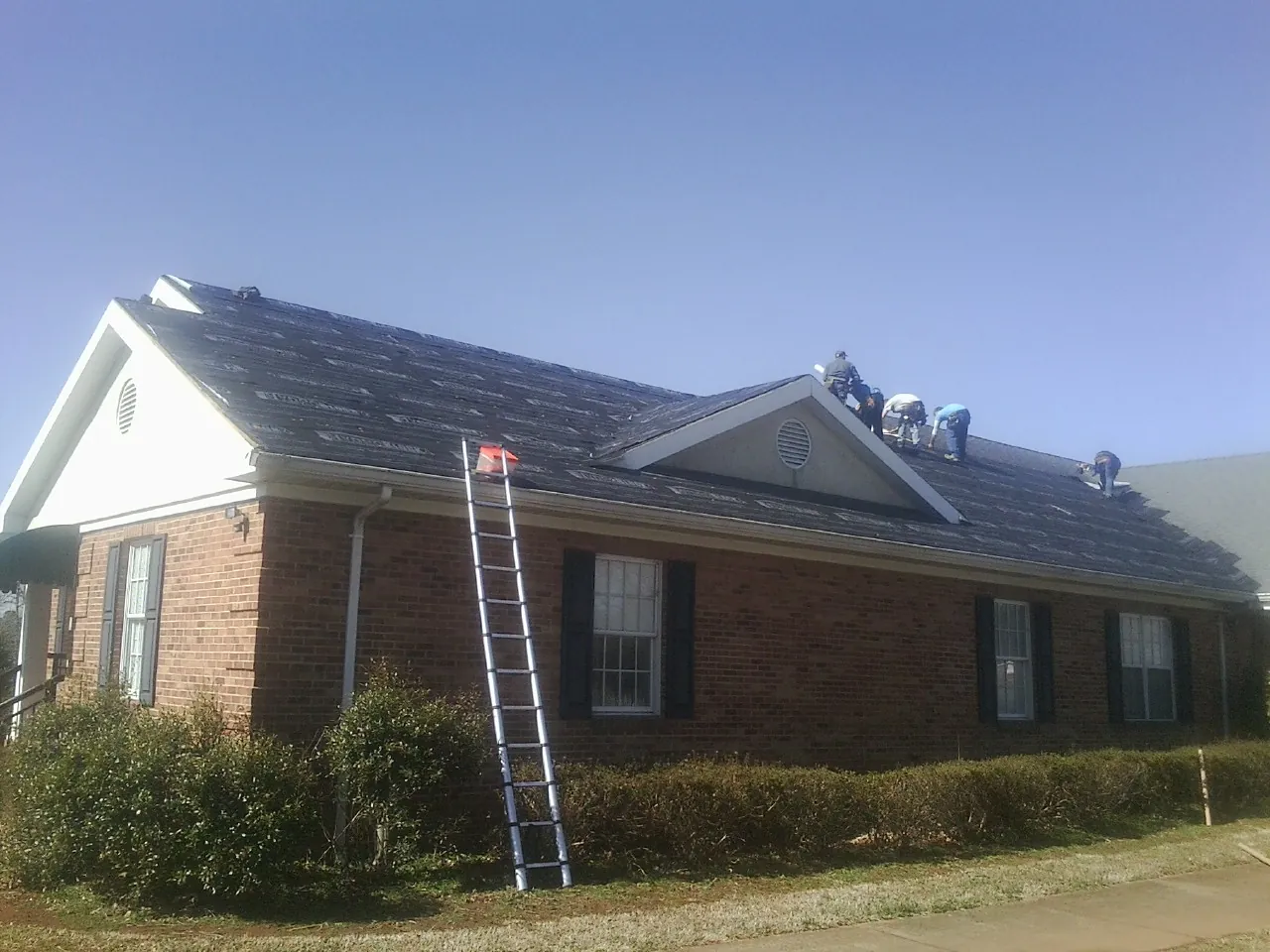 Crew tearing shingles off a brick ranch roof with a ladder set — Metal Roof Repair work by our crew in Greenland