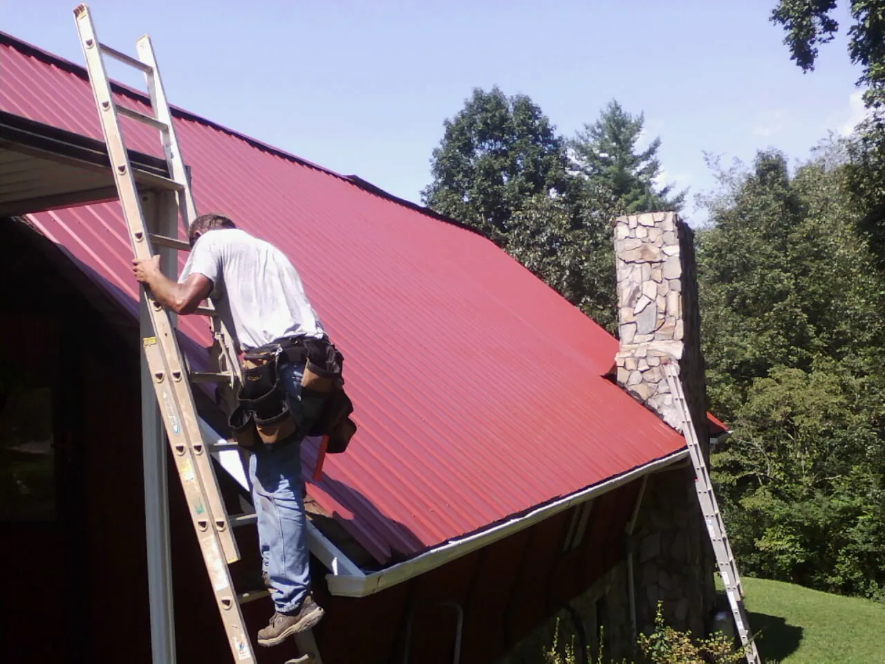 Roofer on a ladder beside a red metal roof and stone chimney — Soffit & Fascia Repair by Roofing Brothers in Greenland